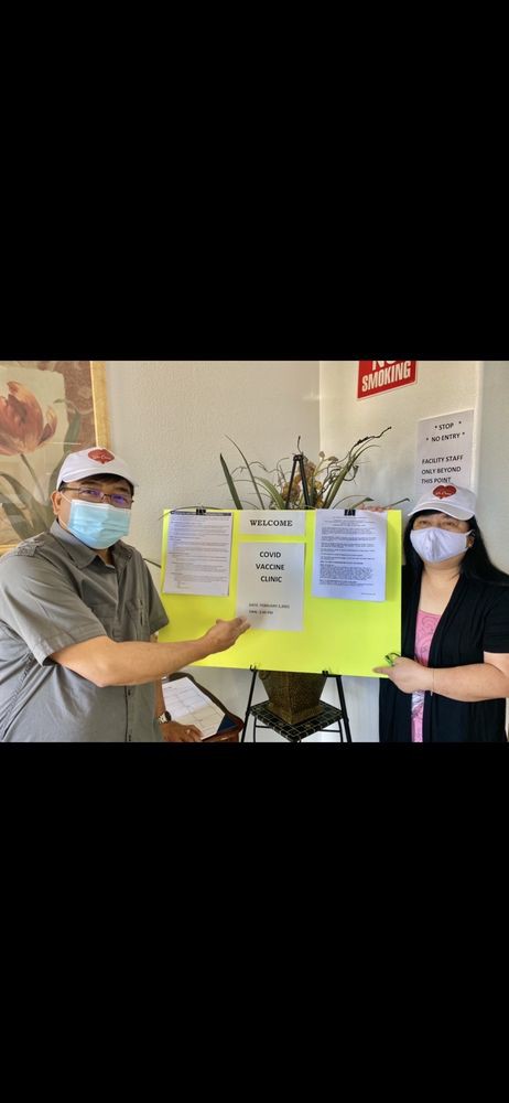 Two individuals wearing masks and hats stand indoors, holding a yellow sign with printed information.