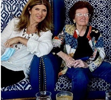 Two women are seated next to each other, smiling and enjoying a relaxed moment indoors.