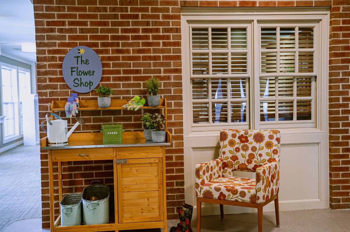 A cozy flower shop corner featuring plants, a colorful chair, and a brick wall backdrop.