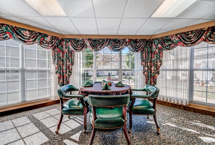 A traditional dining area with a round table and green upholstered chairs, surrounded by large windows and colorful curtains.