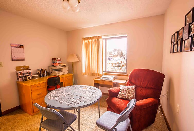 A cozy living room with a round table, armchair, desk, and a window letting in natural light.