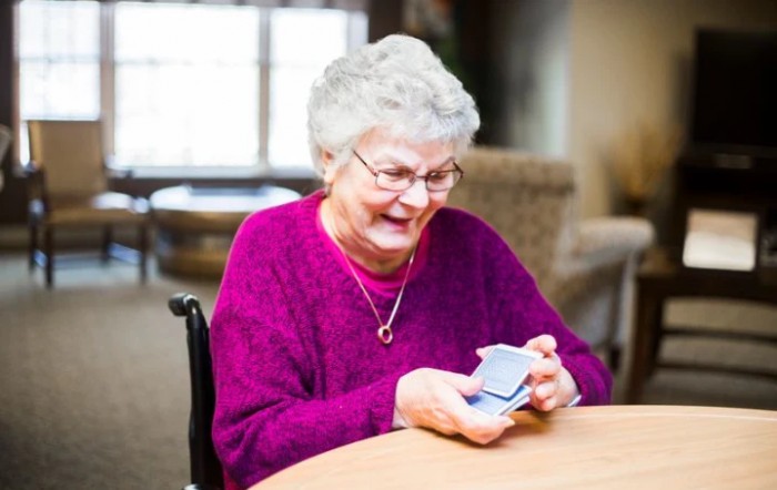 An elderly woman with glasses smiles while playing cards indoors.