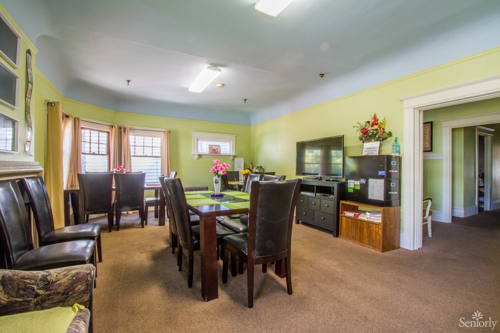 A well-lit dining room with a large table and several chairs, surrounded by windows and decorated shelves.