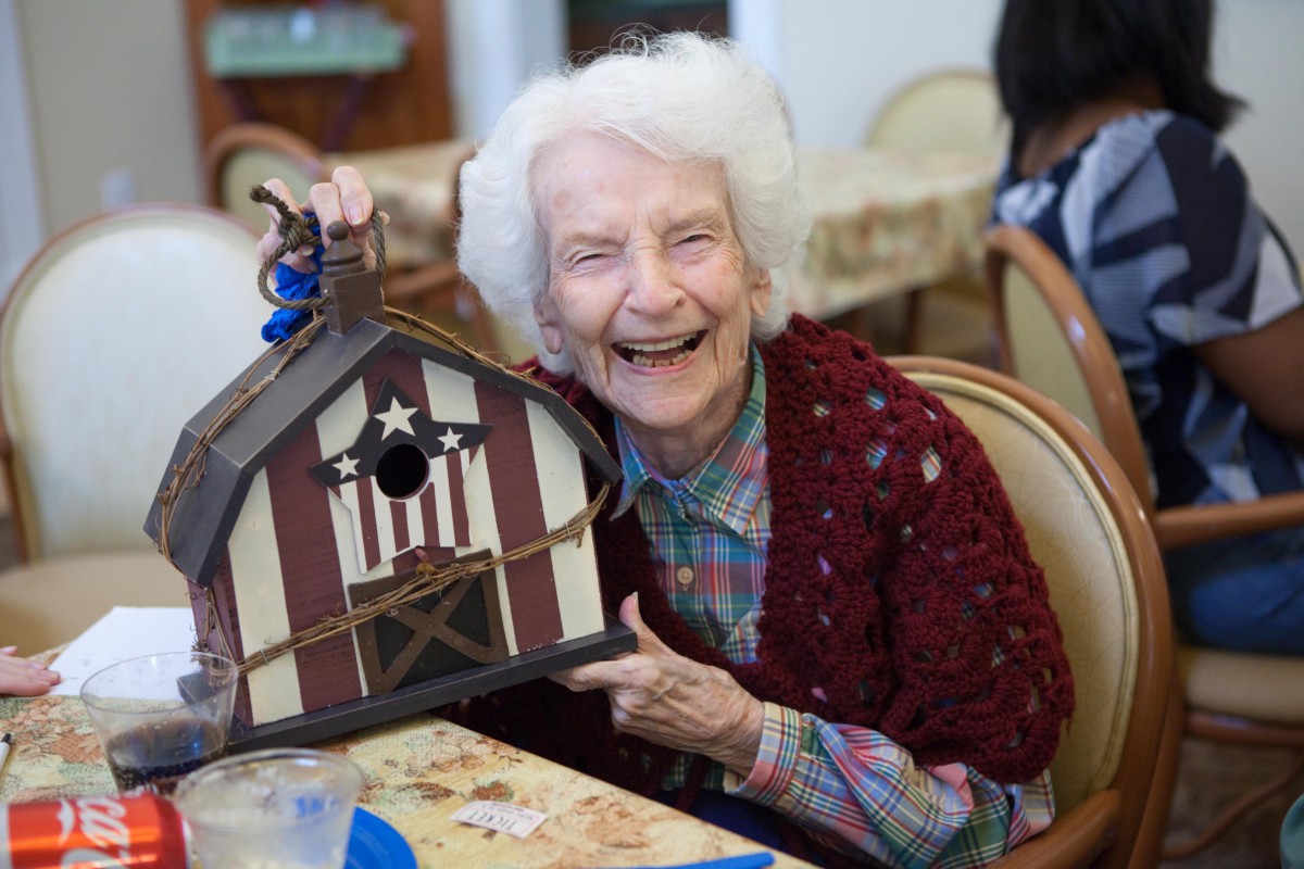 An elderly woman joyfully holds up a handcrafted birdhouse indoors.