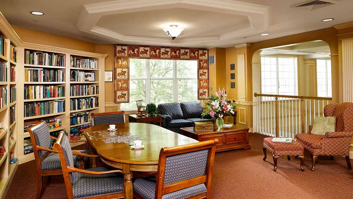 A cozy living room with a library corner, featuring a large table surrounded by chairs, a sofa by the window, and bookshelves filled with books.