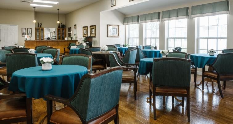 A cozy dining area with blue tablecloths and upholstered chairs, well-lit by large windows.