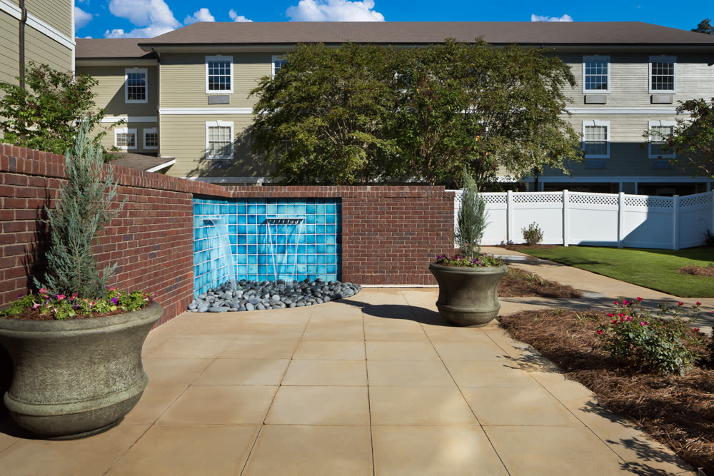 A tranquil outdoor area featuring a small fountain set against a brick wall, surrounded by planters and paved walkways.