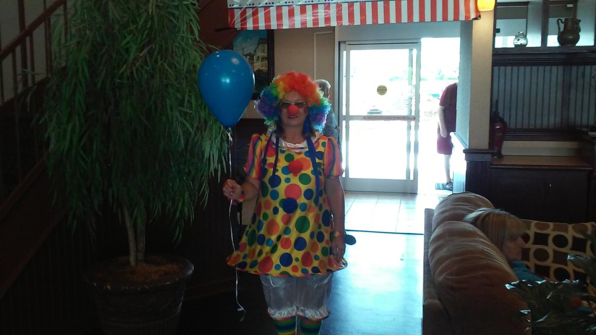 A person dressed as a clown holding a blue balloon indoors.