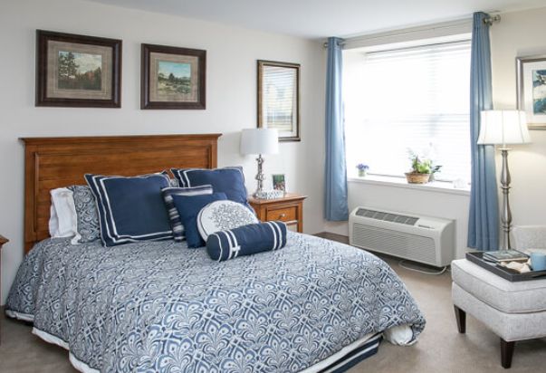 A cozy bedroom featuring blue patterned bedding, framed artwork on the walls, and a sunlit window.