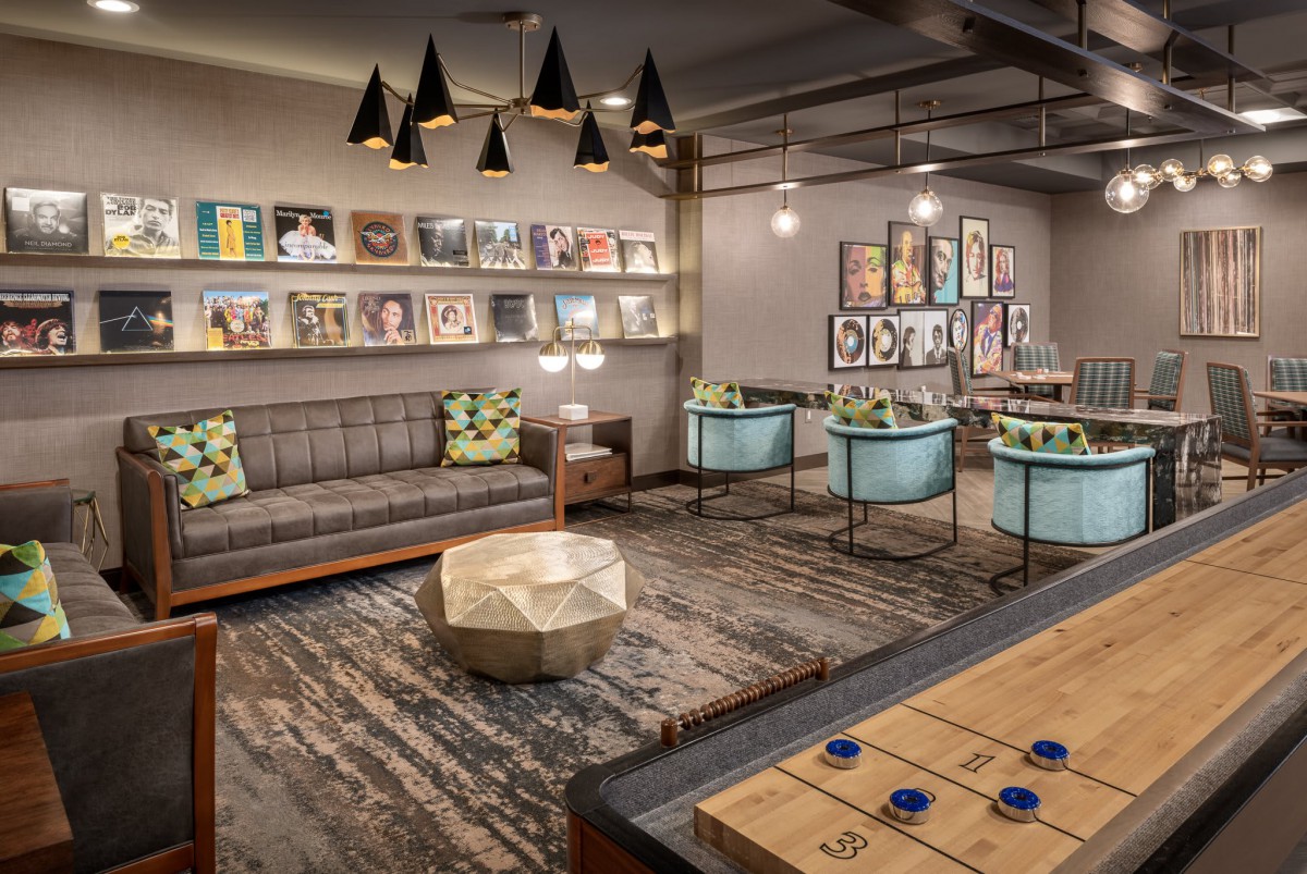 A modern lounge featuring comfortable seating, a bookshelf filled with magazines, stylish artwork on the walls, and a shuffleboard table for entertainment.