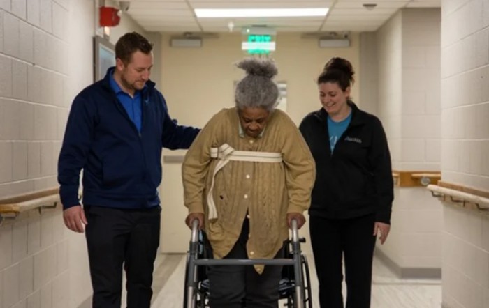 An elderly person using a walker is assisted by two healthcare professionals in a hallway.