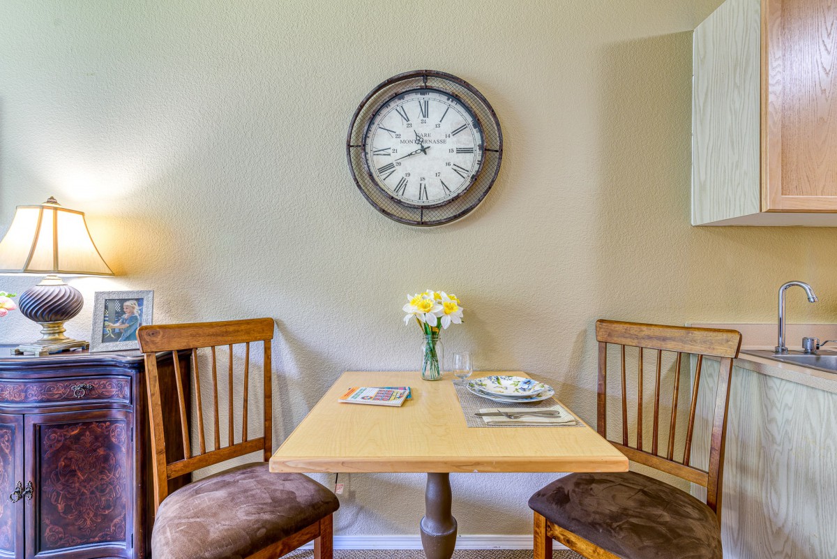 A cozy dining nook with a small table set for two, featuring a wall clock and a vase of flowers.
