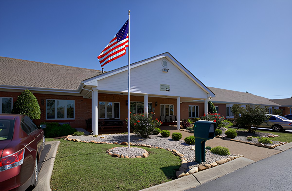 A building with a flagpole displaying the American flag in front of it, surrounded by a landscaped lawn under a clear blue sky.