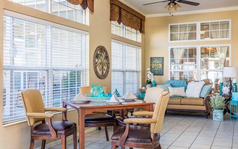 A cozy dining and living area filled with natural light from large windows, featuring a wooden table and chairs surrounded by decorative elements.