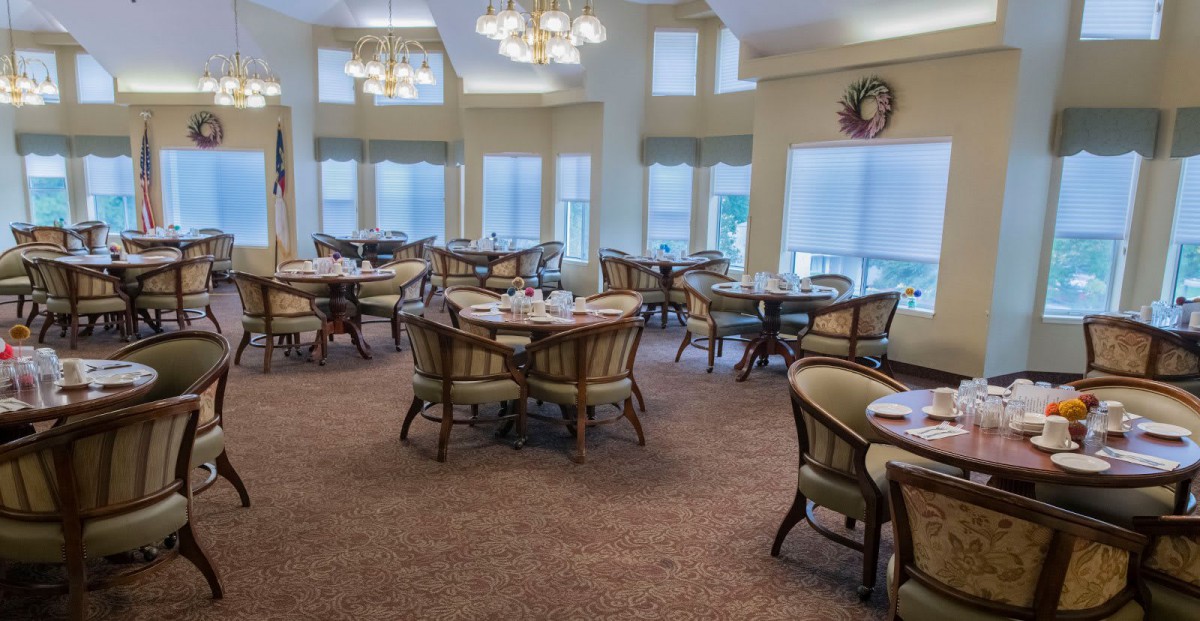A spacious dining room with elegantly arranged tables and chairs, featuring chandeliers and large windows.