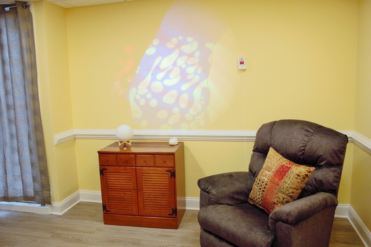 A cozy living room corner with a comfortable armchair beside a wooden cabinet, featuring a colorful projection on the yellow wall.