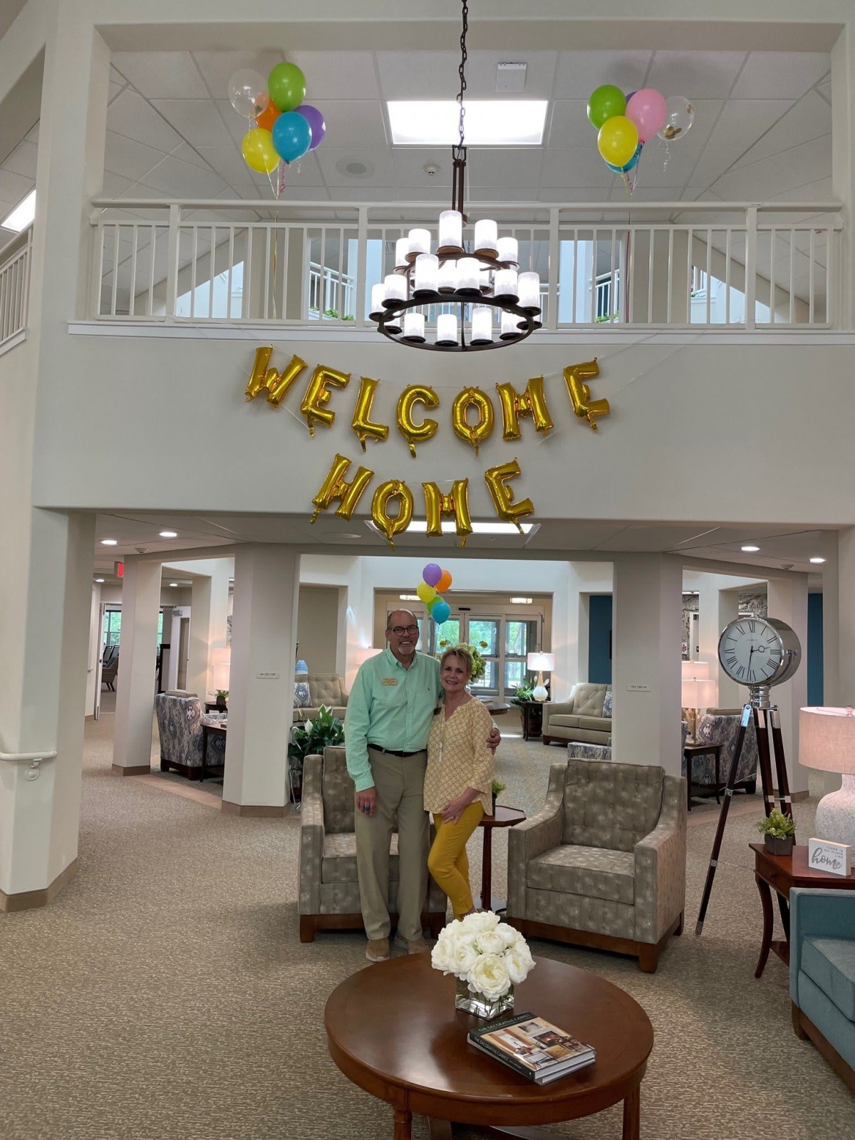 A couple stands in a warmly decorated living room with a 'Welcome Home' banner and balloons.