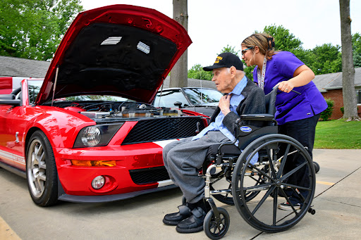 An elderly man in a wheelchair inspects the engine of a red car with the help of a young woman, showcasing community engagement and interest in cars.