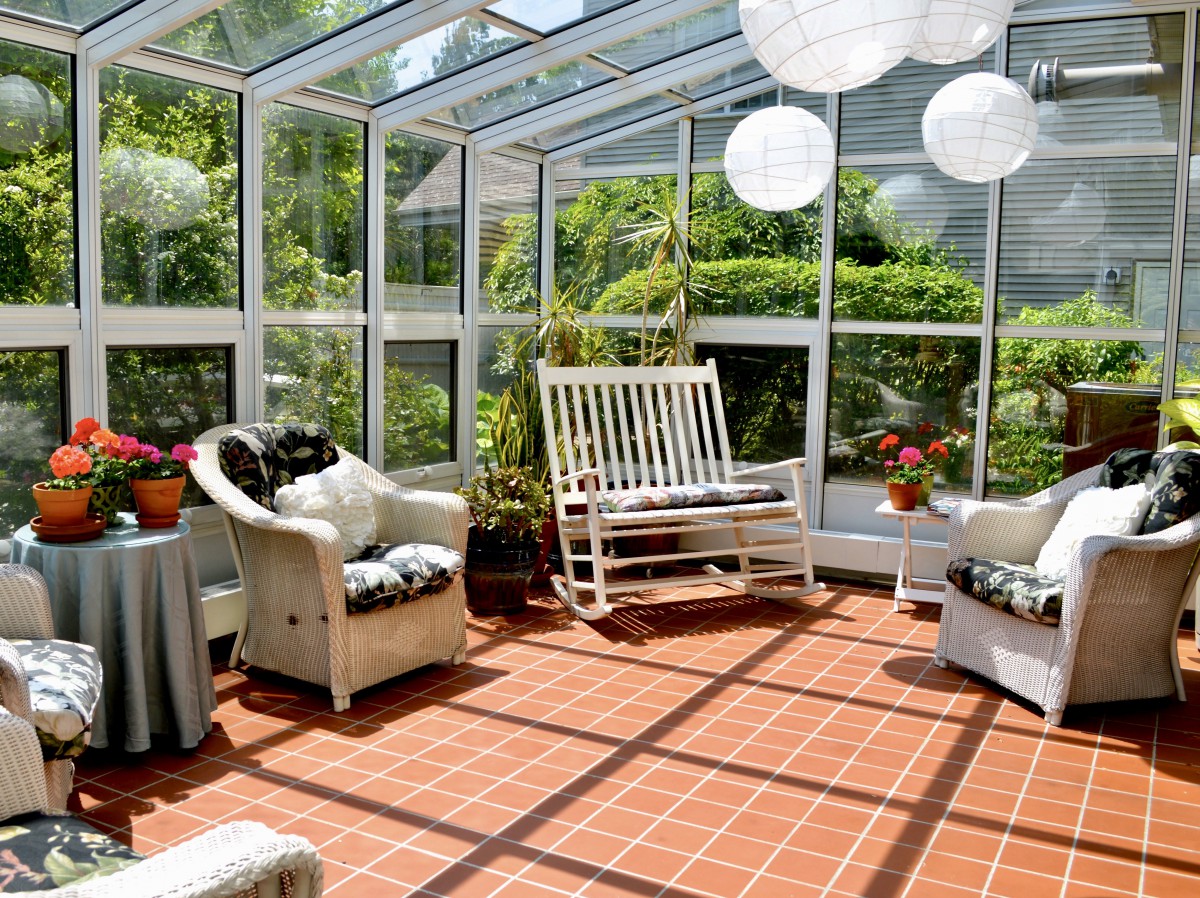 A cozy sunroom with comfortable furniture, surrounded by glass walls, featuring a swing chair and decorative lanterns.