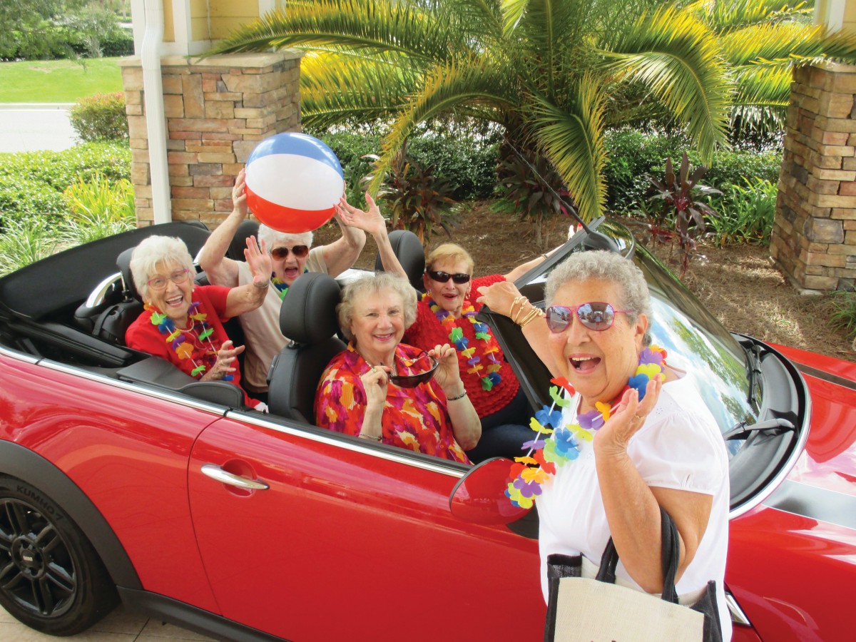 A group of seniors enjoying a vacation in a red convertible, accessorized with tropical shirts and a beach ball.