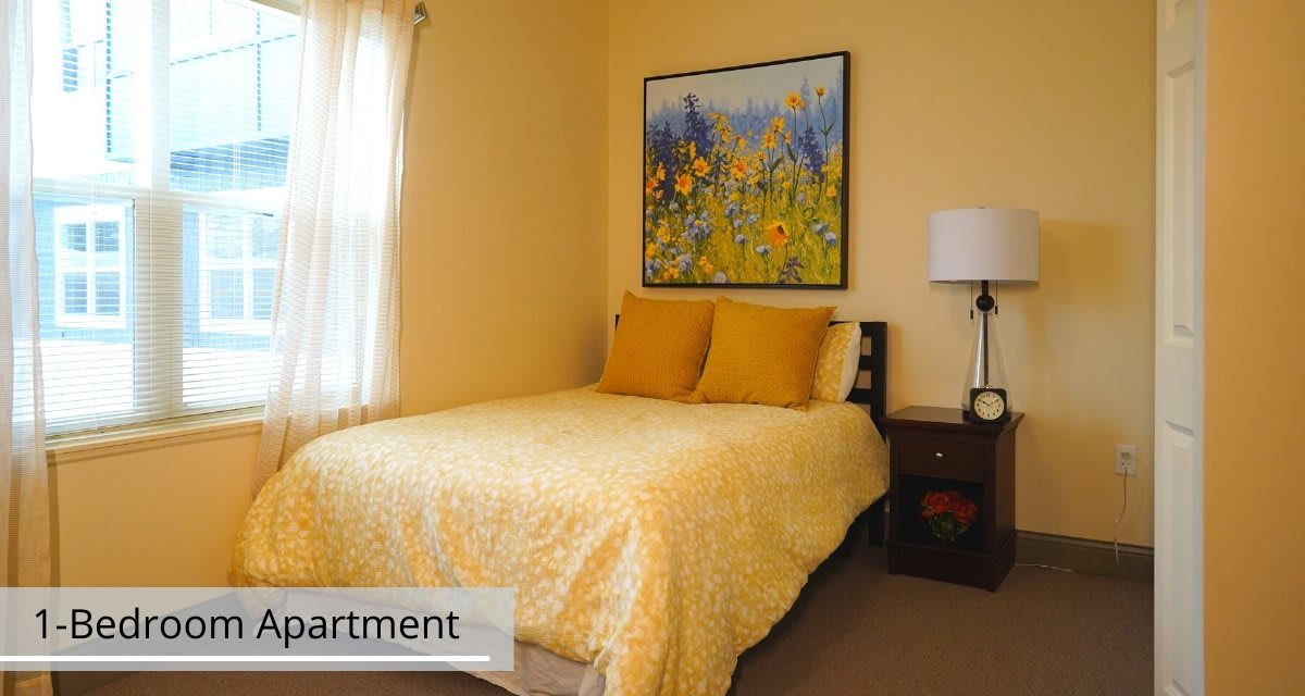 A cozy bedroom with a bed covered in yellow bedding, a floral painting, and a lamp on the nightstand.