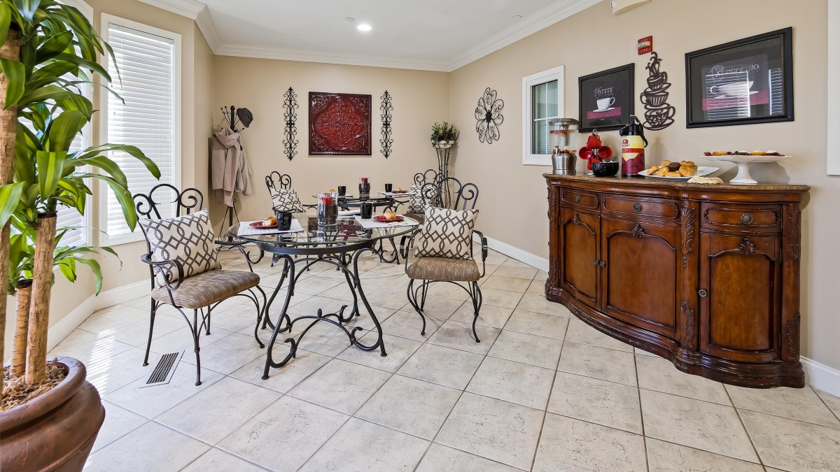 A stylish dining room featuring a glass table and wrought iron chairs set on a tiled floor, with decorative art pieces and a wooden sideboard.