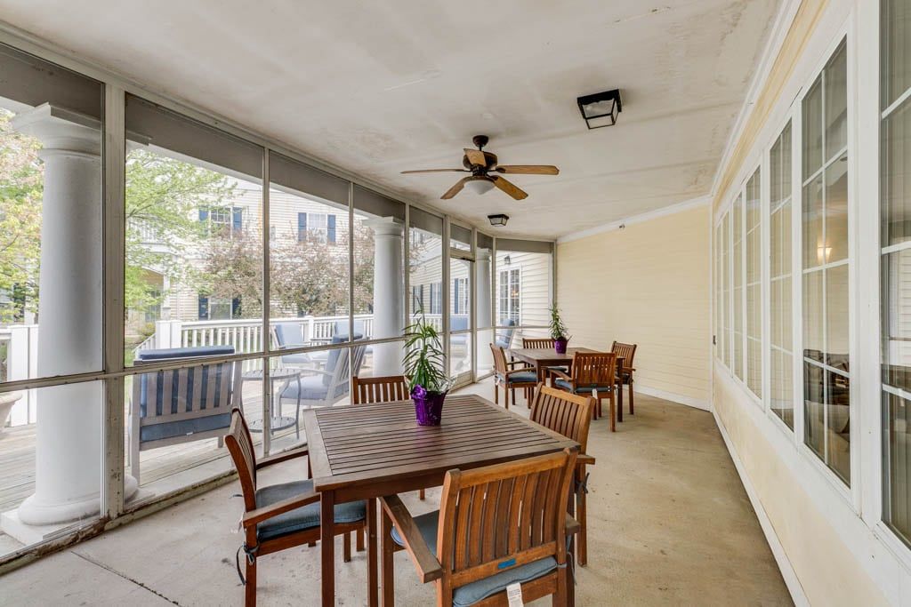 A screened-in porch with outdoor wooden furniture, featuring a ceiling fan and potted plants.