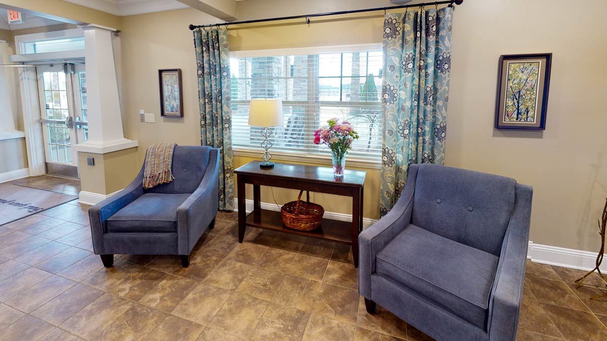 A cozy living room with two blue chairs, a wooden table, and floral curtains by a large window.