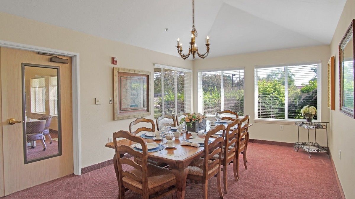 A bright dining room with a large wooden table, surrounded by chairs and illuminated by a classic chandelier.