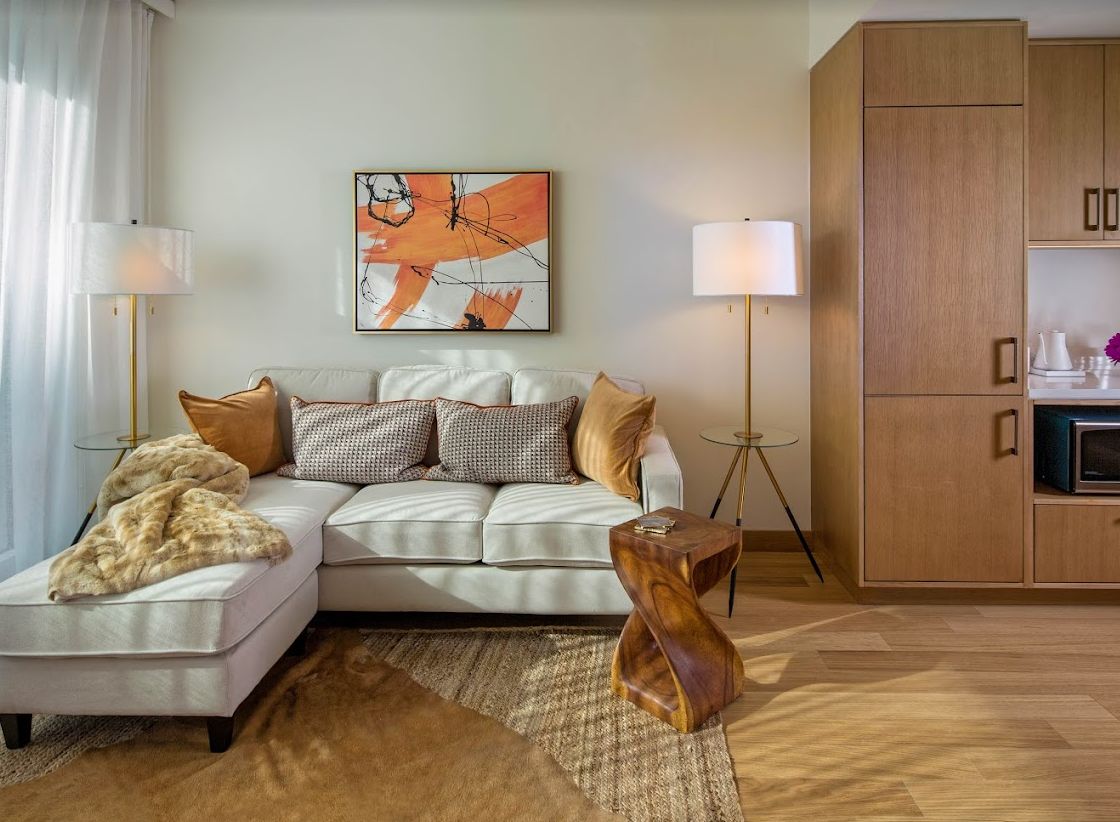 A cozy living room with a white sectional sofa, adorned with cushions, and an abstract painting on the wall. A floor lamp adds warmth to the space, complemented by a wooden side table and a textured rug.