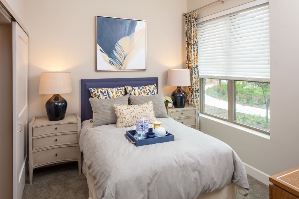 A well-decorated bedroom featuring a neatly made bed with decorative pillows, flanked by two nightstands with lamps, and a piece of abstract artwork above the headboard. Large windows allow natural light to illuminate the room.
