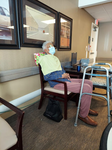An elderly man wearing a mask sits in a waiting area, accompanied by a walker.