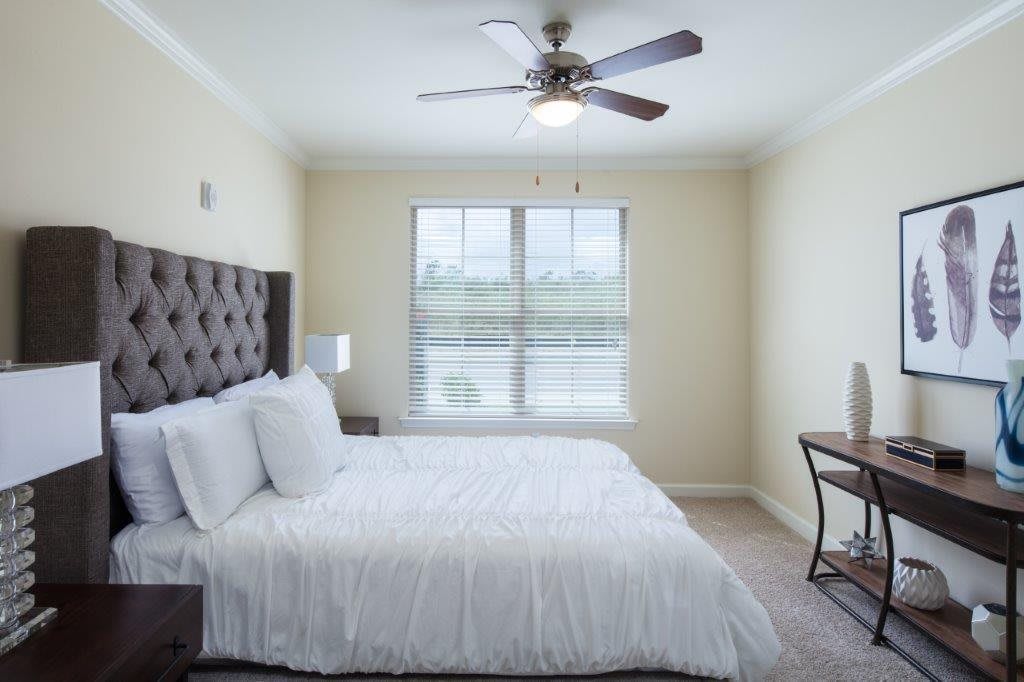 A modern and minimalist bedroom with white bed linens, a ceiling fan, and ample natural light from the window.