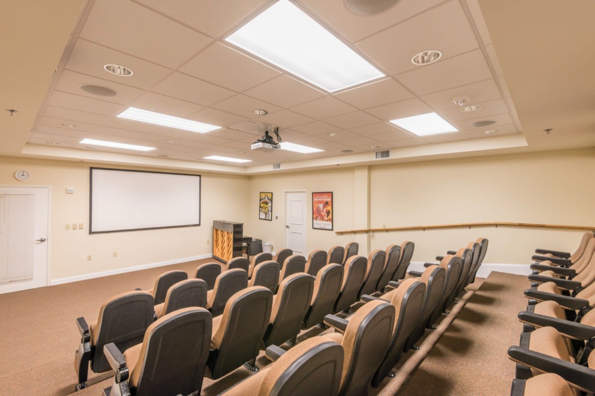 An empty conference room with rows of chairs facing a large screen, equipped with a projector.