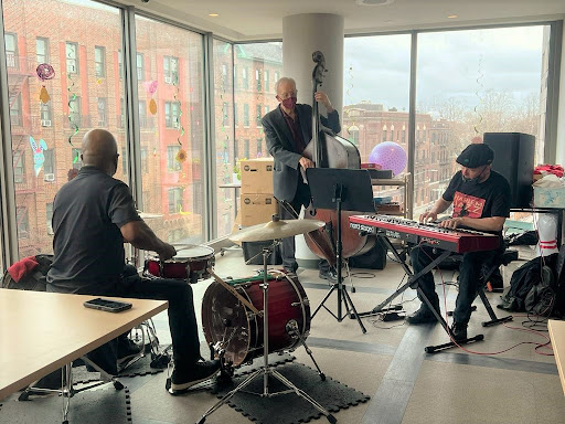 A lively jazz performance featuring a drummer, bassist, and keyboardist in a bright room with large windows.