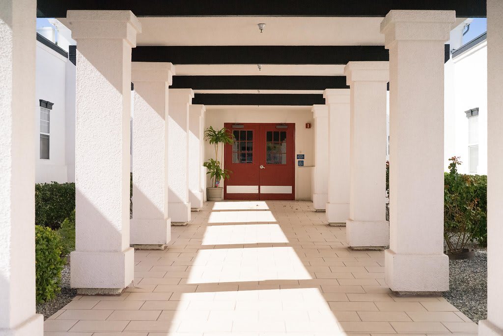 Sunlight casts shadows along a columned corridor leading to a red door.