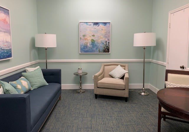 A cozy living room with a sofa, armchair, and two standing lamps, featuring a painting on the wall.