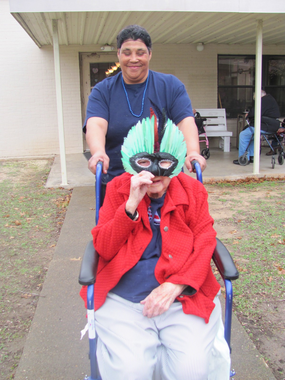 An elderly person wearing a colorful mask is being pushed in a wheelchair by a caregiver outside.