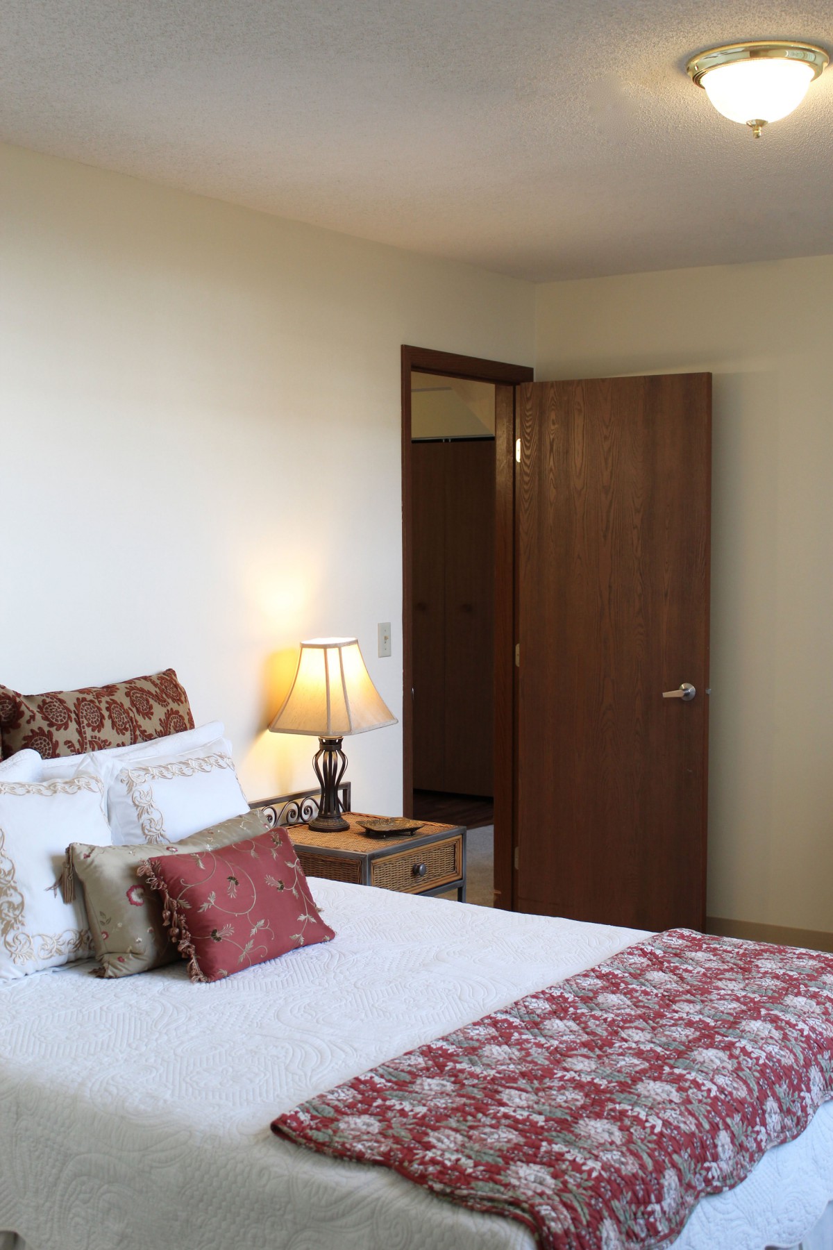 A cozy bedroom with a neatly made bed, decorative pillows, and a warm lamp beside a wooden door.