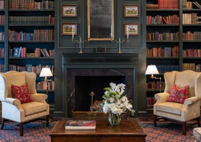 A cozy living room featuring a library with dark green walls, elegant armchairs, and a warm fireplace.