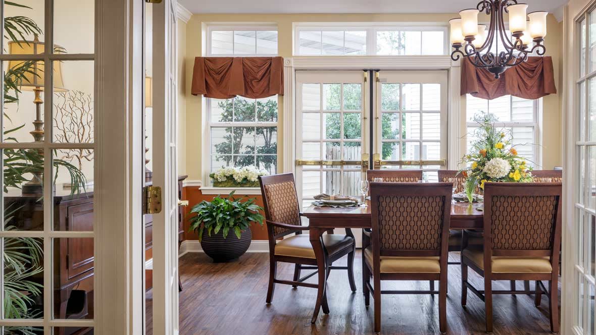 A well-lit dining room with a wooden table and chairs, large windows, and indoor plants.
