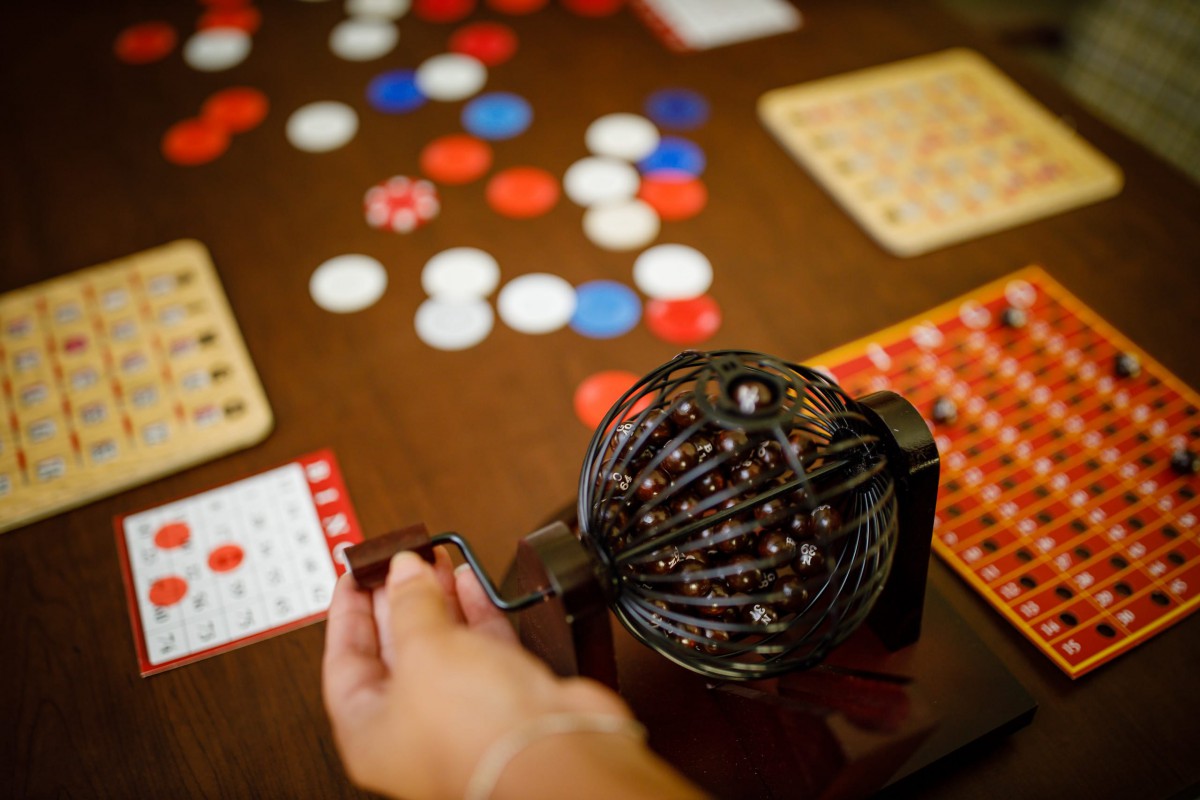A hand turning the handle of a bingo cage, with cards and chips on the table.