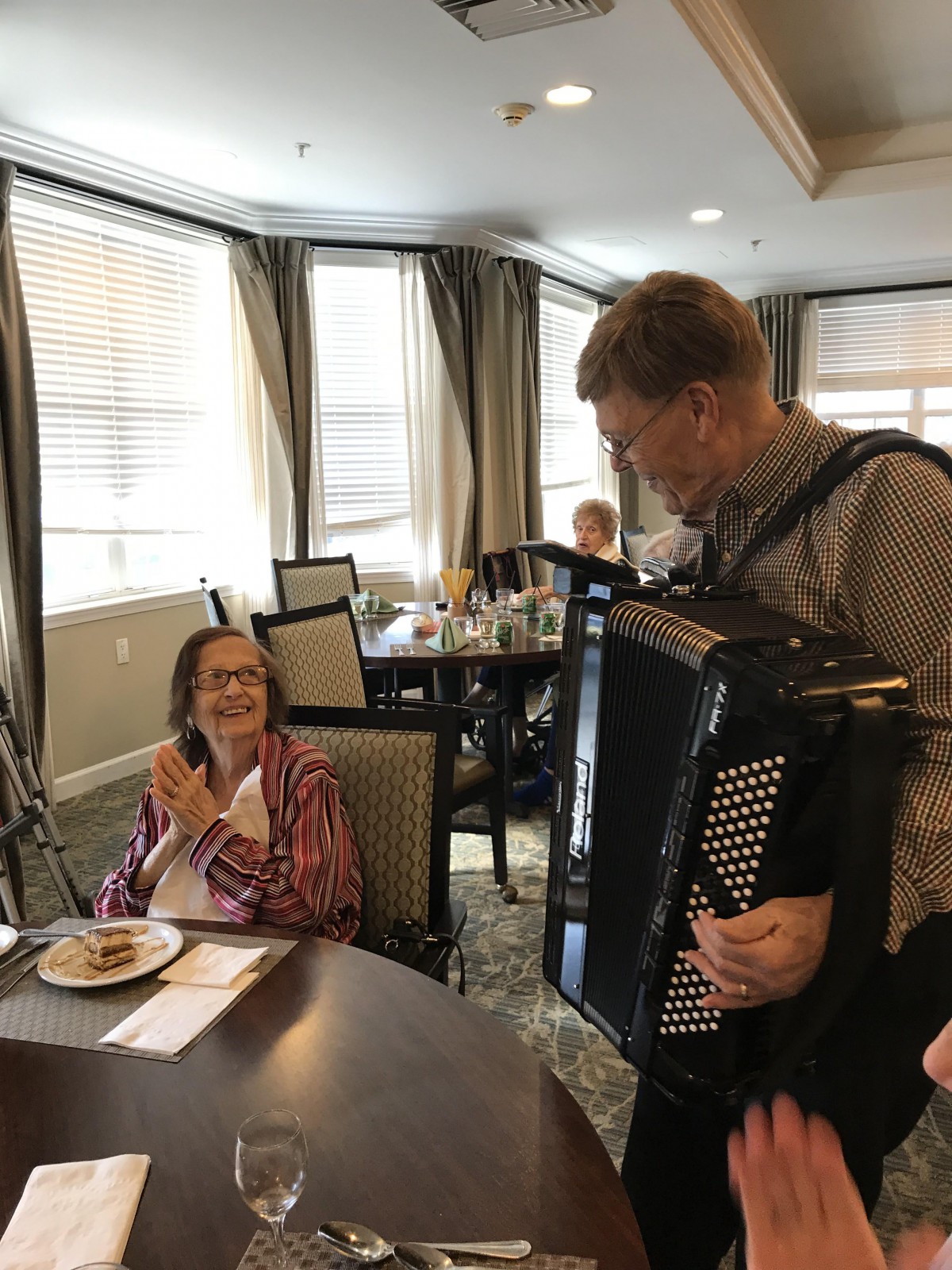 A musician plays the accordion for an elderly woman in a dining room setting.