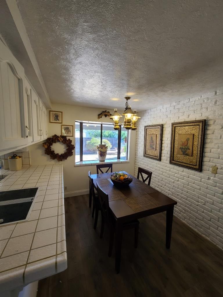 A cozy dining area with a wooden table and chairs, decorated with artwork on white brick walls and illuminated by a chandelier.