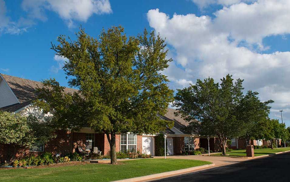 A quiet suburban neighborhood with brick houses and lush green lawns under a partly cloudy sky.