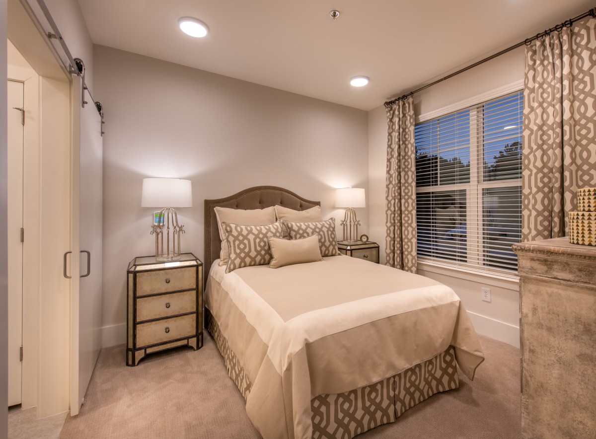 A cozy and elegantly designed bedroom with neutral tones, featuring a neatly arranged bed, bedside tables with lamps, and large windows with drapes.