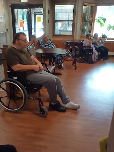 An elderly man seated in a wheelchair, surrounded by other seniors in a care facility.