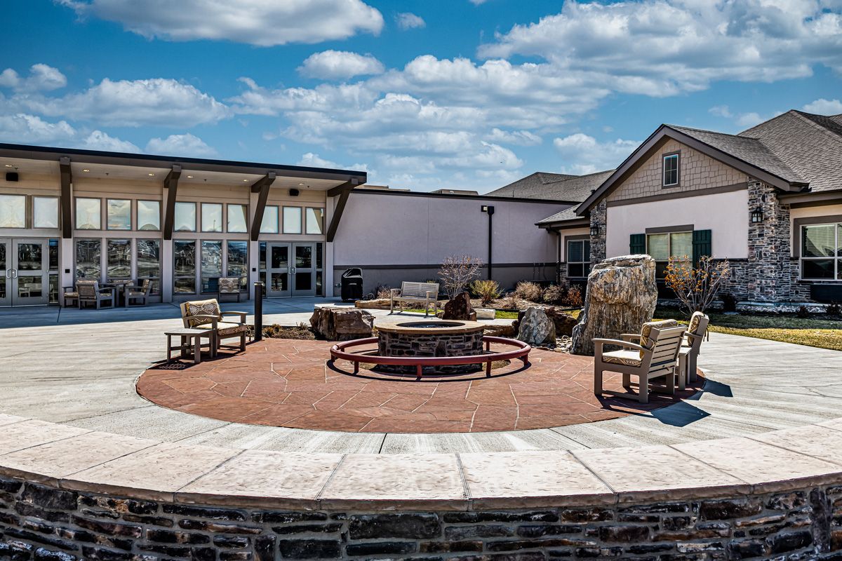 A cozy outdoor patio area featuring a fire pit surrounded by chairs, with buildings and a cloudy sky in the background.