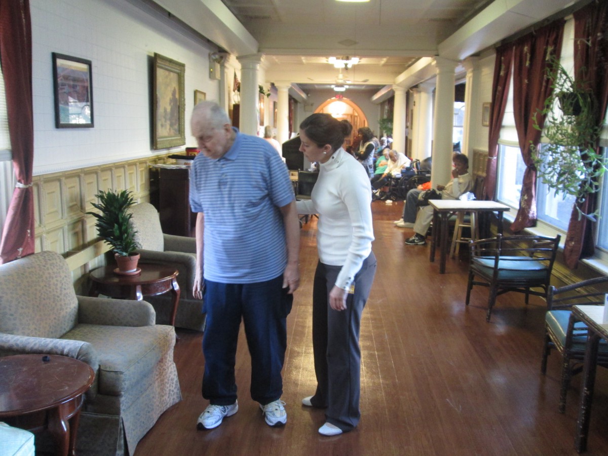 An elderly person receiving assistance in a nursing home hallway.