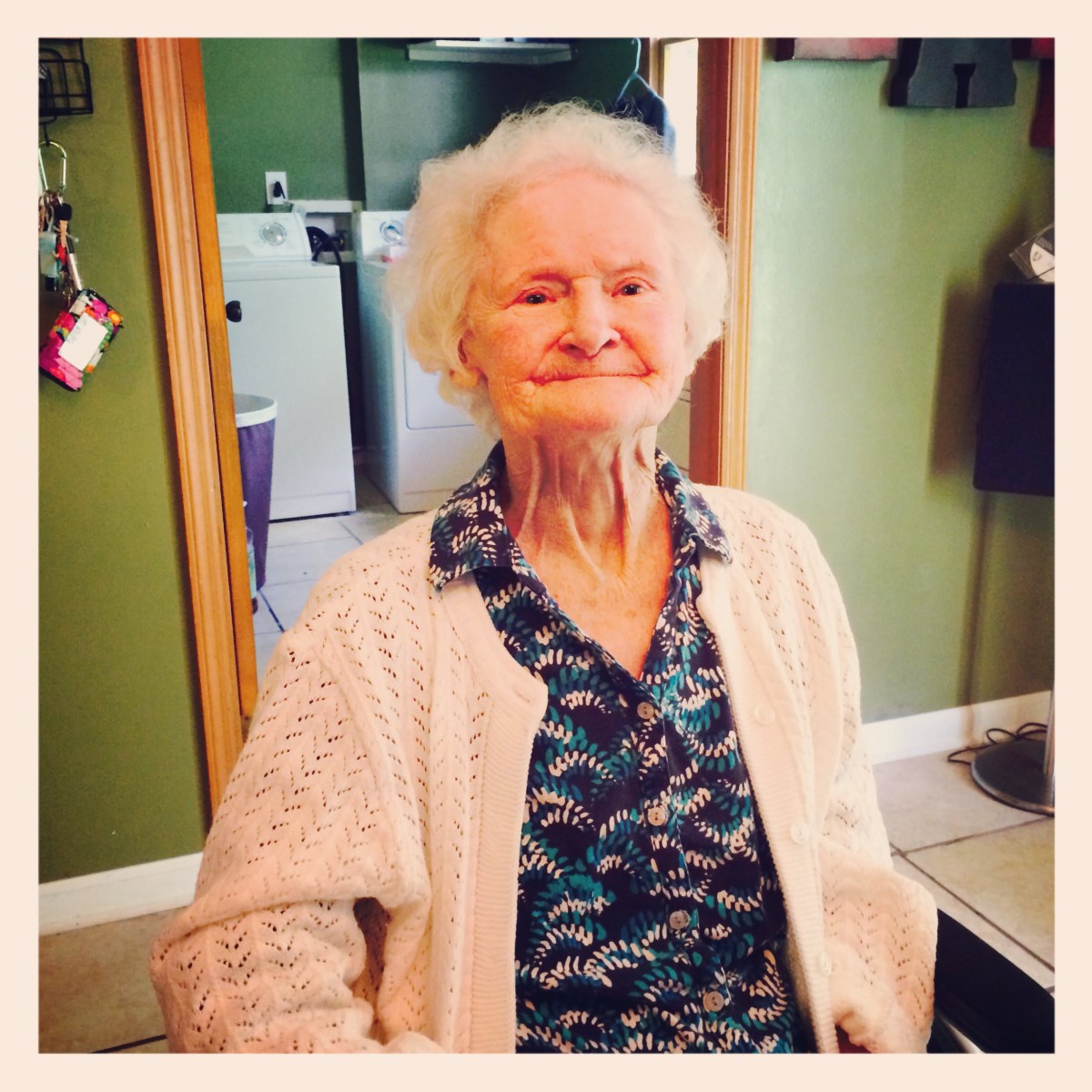 An elderly woman smiling while seated indoors, wearing a cardigan.